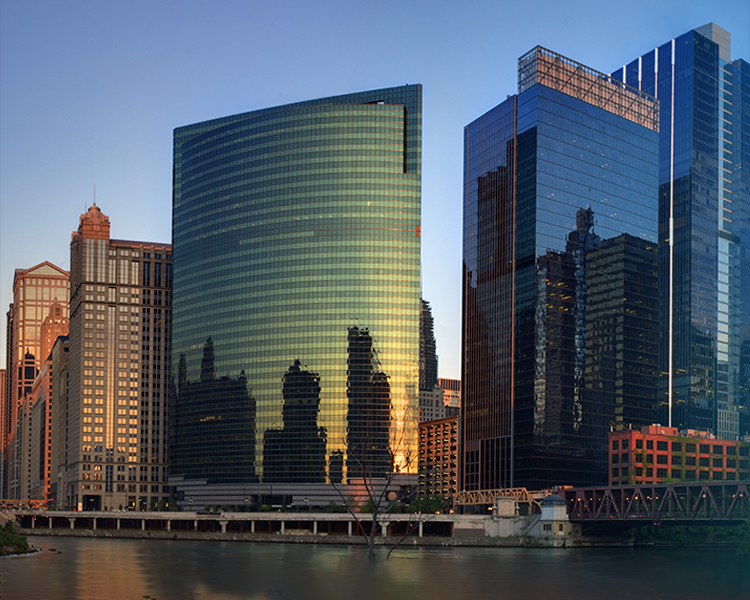 Modern city skyline reflecting the sunset on a riverfront, showcasing a mix of architectural styles.