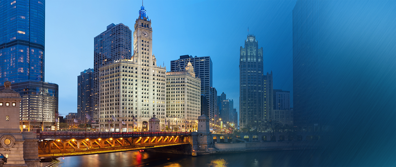 A historic building with a clock tower illuminated at twilight beside a lit bridge over a river, with city lights reflecting in the water.