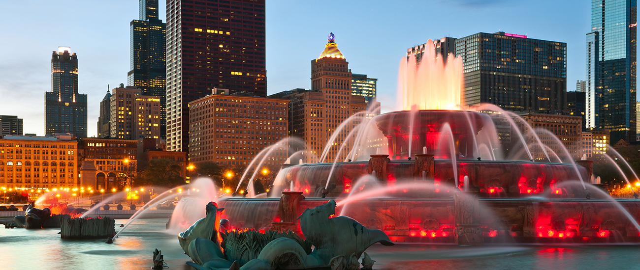 An illuminated fountain with statues at dusk, with the city skyline and a skyscraper with distinctive spires in the background.